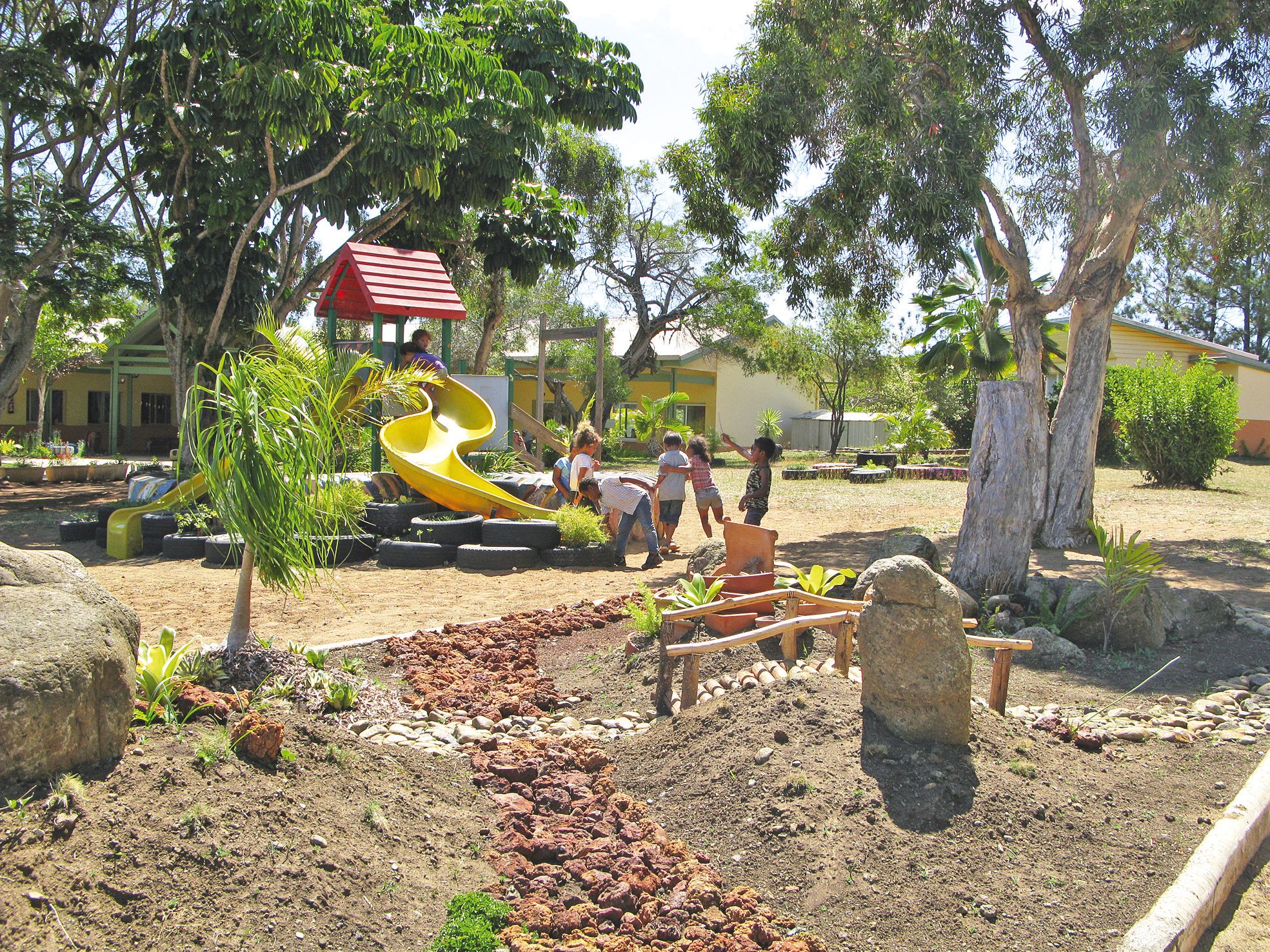 Les parents et le personnel de l’école ont mis en place un jardin japonais dans la cour de l’école maternelle. Les enfants peuvent traverser le petit pont de bois et jouer sur un toboggan entouré de pneus recyclés en jardinières.